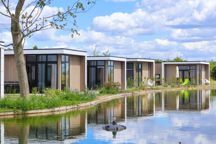 Modern glamping cabins by a tranquil pond in a holiday park, set against a bright sky with lush greenery.