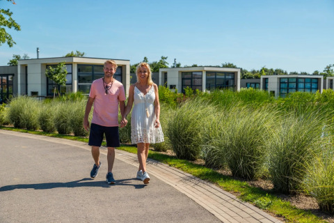 Pareja caminando de la mano en un parque vacacional con cabañas glamping modernas y vegetación alrededor.