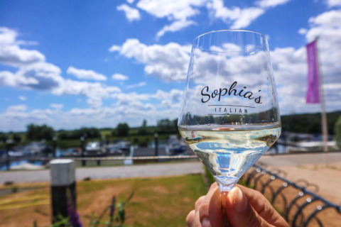 A glass of white wine labeled 'Sophia's Italian' held up outdoors at a holiday park with glamping options.