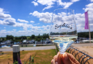 Et glas hvidvin med teksten 'Sophia's Italian' holdes op under en blå himmel ved en luksuscampingplads.
