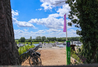 Cykler parkeret ved en flod med både og bådebro i solrigt ferieområde, omgivet af natur og klare skyer.