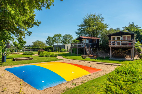 Glamping vakantiedomein met kleurrijke springkussen, boomhutten en groen onder een stralend blauwe hemel.