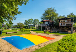 Glamping vakantiepark met kleurrijk springkussen, boomhutten en groene tuinen onder een heldere blauwe lucht.
