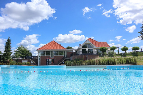 Parque vacacional con glamping, casa moderna de techo rojo y piscina grande bajo un cielo azul.