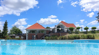Parque vacacional con glamping, casa moderna de techo rojo y piscina grande bajo un cielo azul.