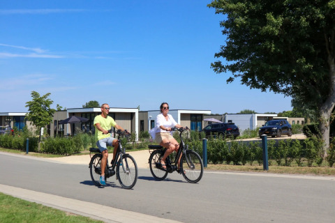 Dos personas montan en bicicleta junto a cabañas glamping en un parque vacacional moderno y soleado.