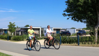 Dos personas montan en bicicleta junto a cabañas glamping en un parque vacacional moderno y soleado.