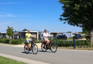 Two people ride bicycles on a sunny day past glamping cabins in a modern holiday park setting.