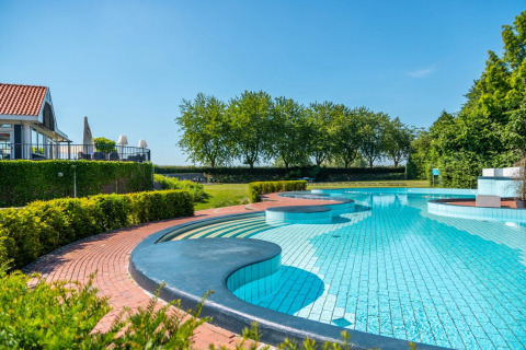 Piscina exterior en parque vacacional con glamping, rodeada de césped y árboles bajo un cielo azul despejado.