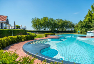 Outdoor swimming pool at a holiday park with glamping, near green hedges and trees under a clear blue sky.