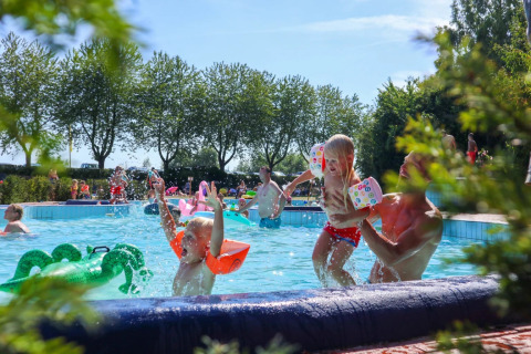 Niños juegan y nadan en una piscina al aire libre en un parque vacacional con glamping, rodeado de árboles y sol.