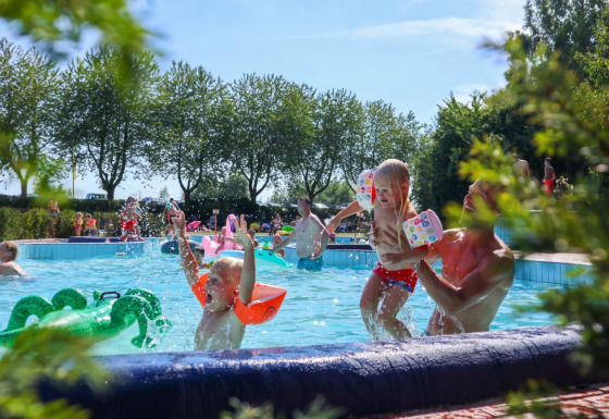 Børn leger og svømmer i udendørs pool på feriepark med glamping, omgivet af træer på en solrig dag.