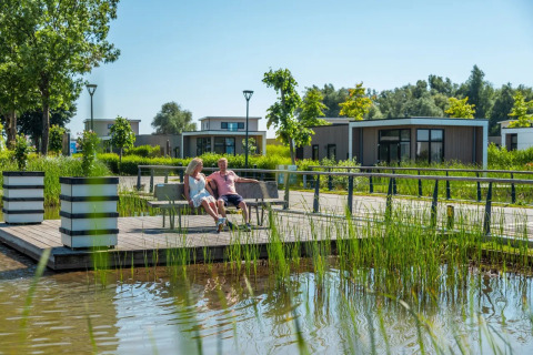 Couple relaxing on a bench by the water at a holiday park, with modern glamping lodges in the background.