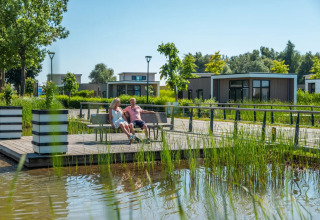Paar entspannt sich auf einer Bank am Wasser in einem modernen Glamping-Ferienpark mit Chalets.