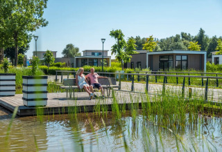 Par slapper af på en bænk ved vandet i en feriepark med moderne glamping-hytter og grøn natur.