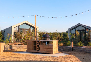 Udendørs scene i feriepark med moderne glamping-hytter, udendørs køkken og belysning om sommeren.