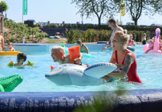 Livredder hjælper et barn med en oppustelig delfin i en pool på en solrig dag i en feriepark.