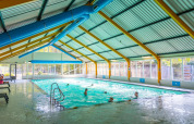 Indendørs swimmingpool med badende gæster på et feriecenter, udsyn til vinduer og grønne omgivelser.