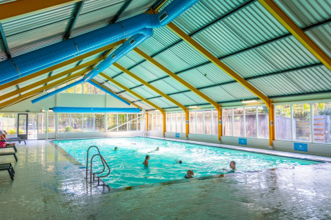 Piscina cubierta con huéspedes nadando en un parque vacacional, ventanas grandes con vista al exterior.