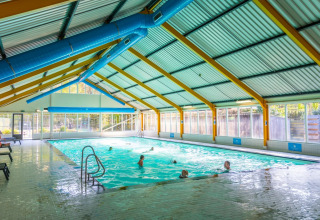 Piscina coperta con ospiti che nuotano in un villaggio vacanze, ampie finestre con vista sul verde.