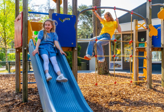 To piger leger på legeplads med rutsjebane og klatrestativ i en feriepark med glampingfaciliteter.