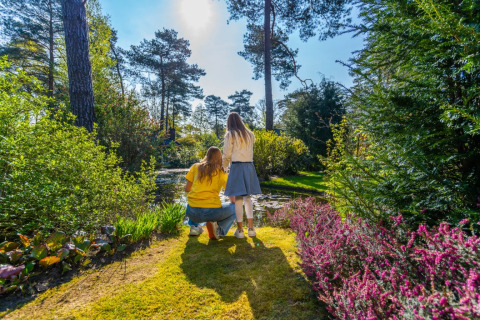 To personer nyder naturen ved en dam i en feriepark med glamping, omgivet af blomster og solskin.