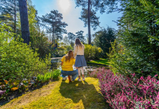 Twee mensen genieten van de natuur aan een vijver in een glamping-vakantiepark, omringd door bloemen.