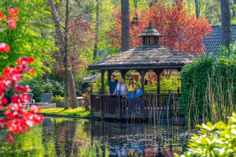 Familia disfruta en un cenador junto a un estanque, rodeados de coloridos árboles en un parque de glamping.