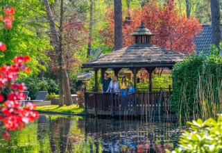 Familie nyder en hyggelig stund i et lysthus ved en dam i en grøn og farverig feriepark til glamping.