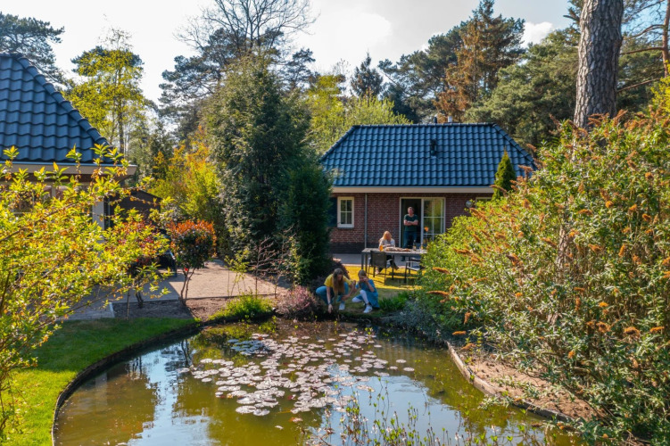 Family relaxing at a pond in front of cozy glamping lodges surrounded by trees at a holiday park.