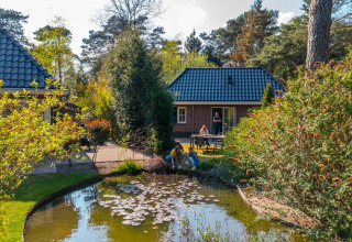 Familie nyder ferie ved dam foran hyggelige glamping-hytter i en grøn feriepark med træer og natur.