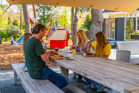 Gezin eet buiten aan picknicktafel in een vakantiepark met speeltuin en glampingaccommodaties.