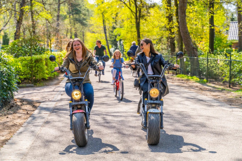 Two women laughing while riding electric scooters on a sunny park path with children cycling behind them.