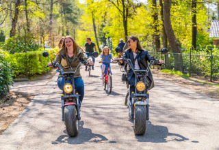 Twee vrouwen rijden lachend op elektrische scooters door het vakantiepark, met kinderen op fietsen achter.