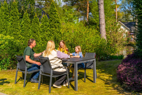 En familie nyder en solrig eftermiddag ved et bord i en grøn have på en feriepark med glamping i baggrunden.