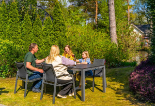 Een gezin zit samen aan een tuintafel in de zon bij een vakantiepark met glampingaccommodaties op de achtergrond.