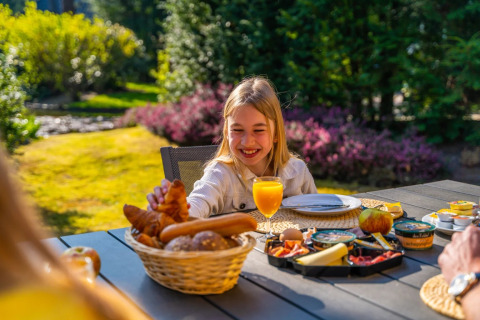 Niña desayunando al aire libre con croissants y zumo en un parque vacacional con glamping.