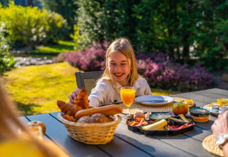 Niña desayunando al aire libre con croissants y zumo en un parque vacacional con glamping.