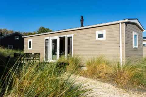 Lodge moderno de color beige con ventanas grandes y mesa exterior, rodeado de césped y cielo azul claro.