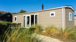 Lodge moderno de color beige con ventanas grandes y mesa exterior, rodeado de césped y cielo azul claro.