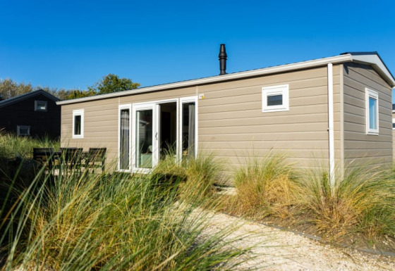 Lodge moderno de color beige con ventanas grandes y mesa exterior, rodeado de césped y cielo azul claro.
