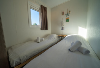 Ein einfaches Lodge-Zimmer mit zwei Einzelbetten, weißen Laken und gerollten Handtüchern am Fenster.