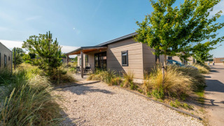 Lodge moderna con fachada de madera, camino de grava y vegetación bajo un cielo soleado y azul.
