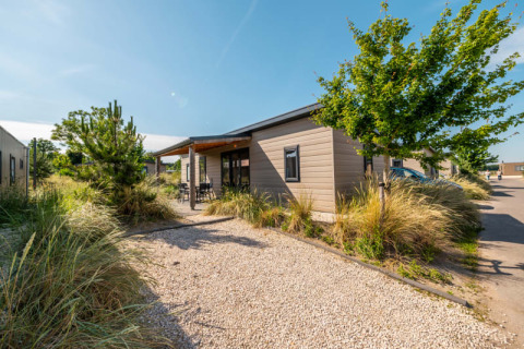 Moderne lodge med træfacade omgivet af grønne planter og et grusbelagt stisystem under blå himmel.