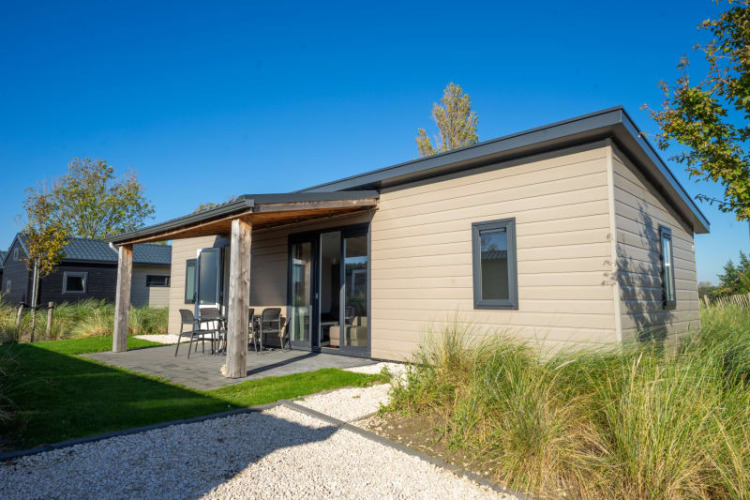 Photo of a modern lodge with a covered patio, outdoor furniture, and greenery under a clear blue sky.