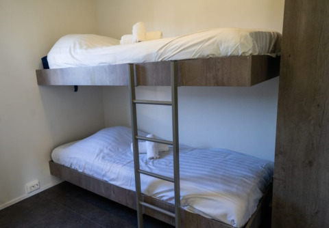 Værelse på en lodge med køjesenge i træ, hvide lagner og håndklæder på hver seng, enkelt og hyggeligt.