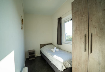 Petite chambre simple lumineuse dans un lodge avec lit simple, fenêtre, armoire et table de chevet.