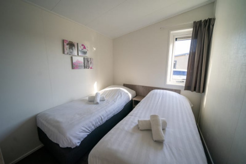 Chambre lumineuse dans un lodge avec deux lits simples, fenêtre, rideaux gris et tableaux muraux.