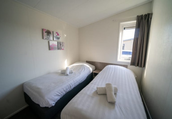 Chambre lumineuse dans un lodge avec deux lits simples, fenêtre, rideaux gris et tableaux muraux.