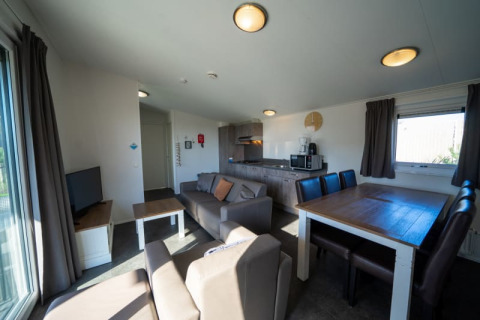 Binnenzicht van een gezellige lodge met zetel, eettafel, kitchenette en ramen met gordijnen.